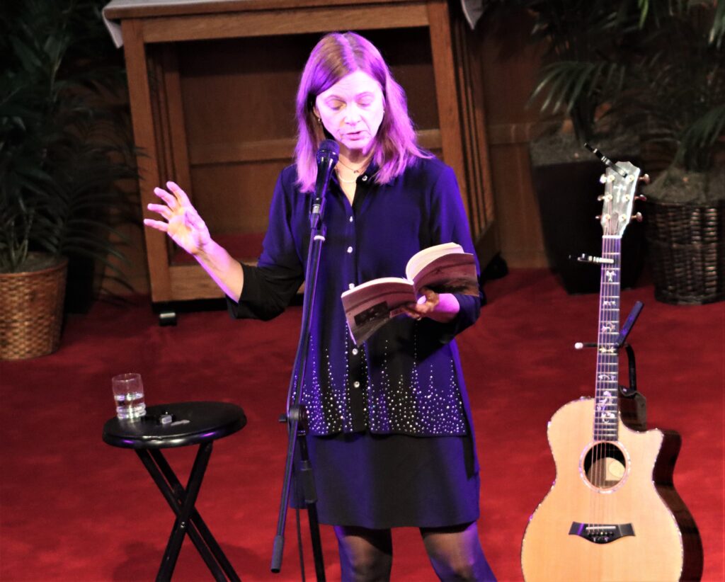 Picture of a middleaged woman (Carrie Newcomer) holding and reading from a book, standing next to a guitar.