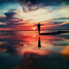Image of a colorful sunset over water and a person standing with arms outstretched.