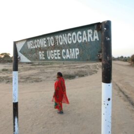 Sign that says, "Welcome to Tongogara Refugee Camp." A woman wearing a red dress stands on sandy ground.