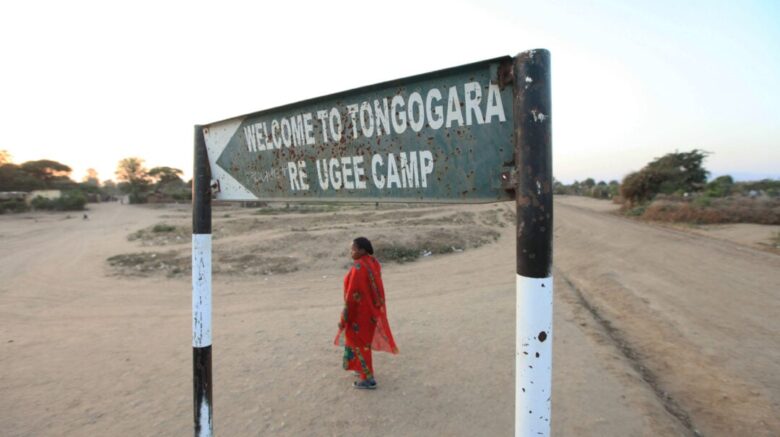 Sign that says, "Welcome to Tongogara Refugee Camp." A woman wearing a red dress stands on sandy ground.